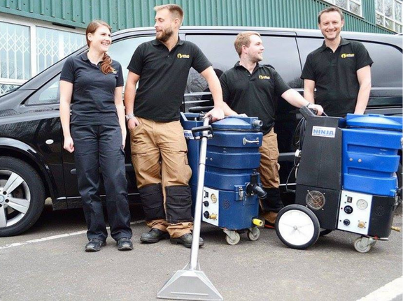 This is a photo of Beddington Carpet Cleaning carpet cleaners (three men and one woman) standing in fromt of their black van, with two steam cleaning carpet machines next to them